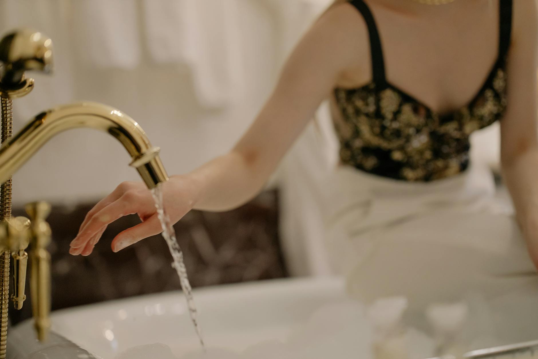 An elegant woman washes her hand under a golden faucet indoors.