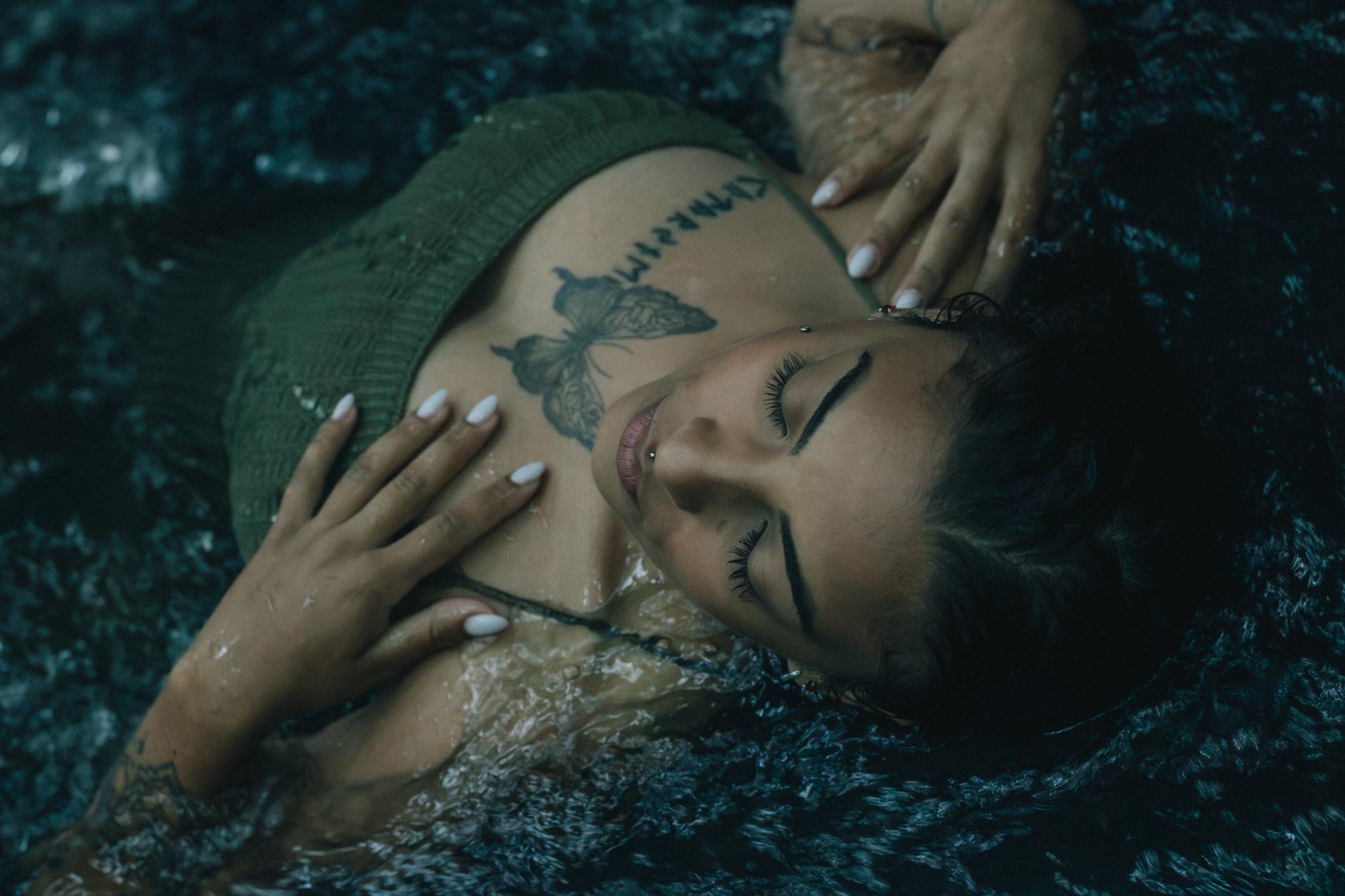 A peaceful woman with a tattoo relaxes in dark water, eyes closed.