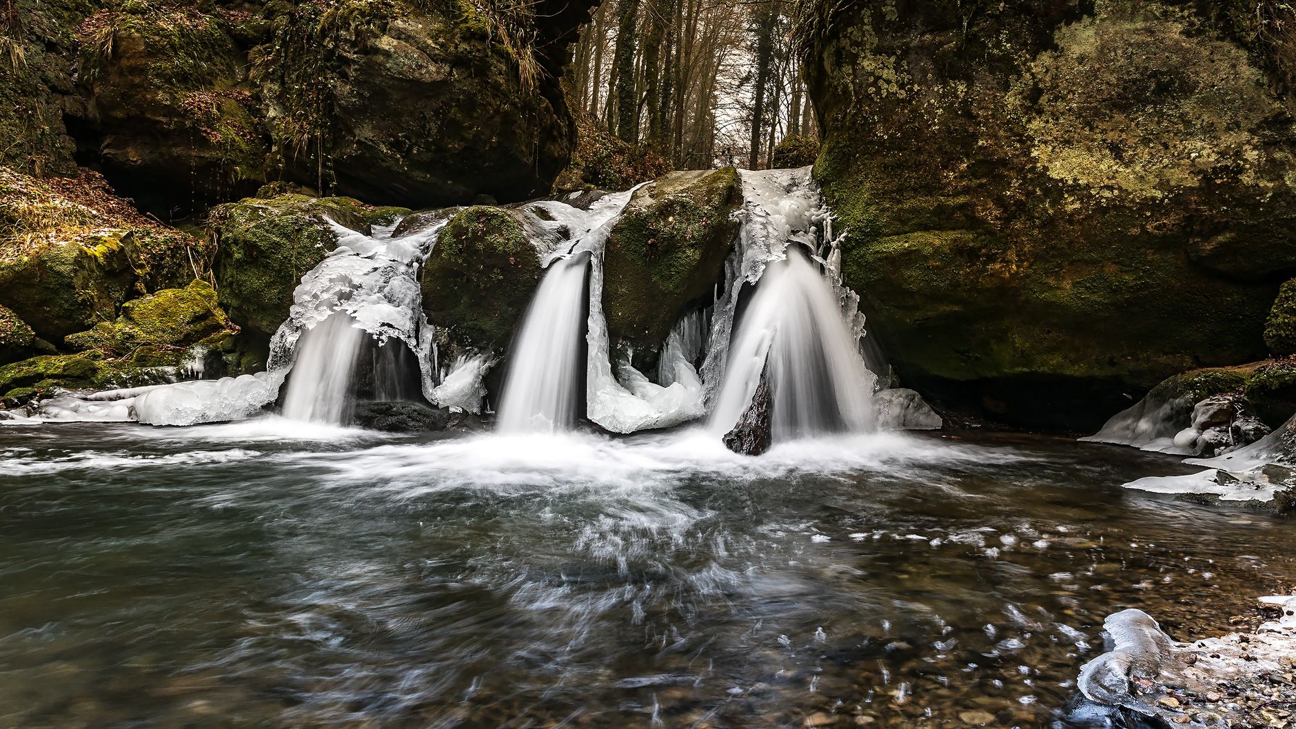 A serene frozen waterfall cascading over moss-covered rocks in a tranquil forest setting.