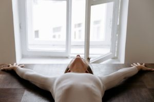Elegant woman in a white bodysuit stretches with arms apart near an open window, capturing serenity indoors.