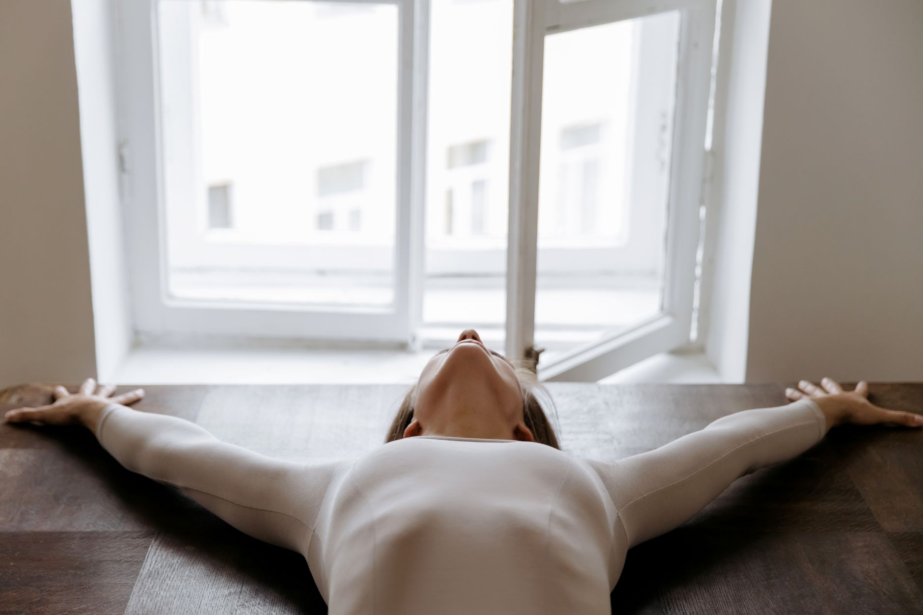 Elegant woman in a white bodysuit stretches with arms apart near an open window, capturing serenity indoors.